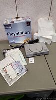 Sony PlayStation console with cables, manuals, and original Dual Shock box displayed on a table