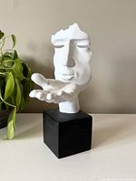 Front view of the white blowing kiss sculpture with stylized human face and hand extended on black cube base, next to plants.