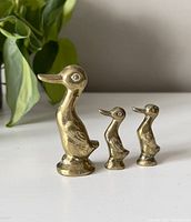 Three vintage brass duck figurines displayed side by side on a white surface with a plant in background.