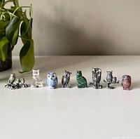Photo showing all 8 miniature owl figurines in a row, displayed on a white surface with a plant leaf in the background. Various materials visible including metal, glass, marble, and ceramic.