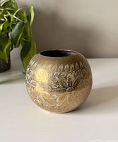 Front and side view of the rounded vintage brass vase showing embossed floral and leaf decoration with warm aged patina on a white surface and houseplant in background.