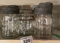 Four antique canning jars displayed side by side on a shelf, from brands Ball, Gem, Jewel, and others. Metal lids visible, some showing rust.