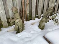 Two weathered concrete statues outdoors partially covered in snow near a wooden fence. Both are children figures holding baskets.