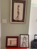 Three framed artworks displayed on a wall and floor: one monochrome sketch in black frame and two colorful prints in wood frames.