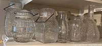Overview of thirteen clear pressed glass jars and bottles on shelf, showing varied patterns, shapes and metal handles.