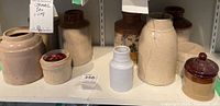 Shelf holding eight vintage ceramic crocks and jars ranging from small to medium size, mainly beige with some brown-glazed tops, showing chips, cracks and wear.