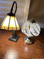 Photo of two table lamps, one with amber stained glass shade and bronze base, the other with frosted floral glass shade and gold plated round base.