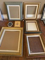 Seven assorted picture frames in various sizes and finishes arranged on wooden floor.