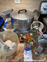 Overview photo showing aluminum cake storage container, plastic mixing bowl with beaters, metal mesh sifter, measuring cups, and other kitchen tools on stone countertop.