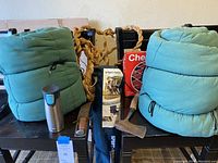 Two green sleeping bags rolled up, black SportsBrella chair with wooden armrests, boxed checkers set, rope, and a Contigo water bottle on a black table.
