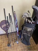 Golf clubs in bag with extra clubs and Prince tennis racket cover leaning against wall on brown tile floor.