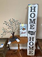 Three items: metal tree sculpture with curled green leaves on base, smaller rustic wooden sign with 'Home Sweet Home' and decorative flower, and large distressed white wooden sign reading 'HOME sweet HOME'.