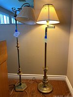 Both vintage floor lamps standing side by side, one lit showing working condition, both have fabric shades and metal bases with ornate details.