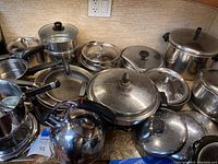 Photo showing collection of various pots, pans, kettle and lids arranged on countertop, highlighting black handles and metallic finishes.