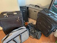 Several pieces of luggage including large black suitcases with wheels, a hard-sided black briefcase style case, a grey textured bag with black handles, and a black duffel bag. Items show dust and some wear.