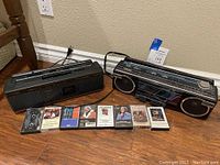 Both Toshiba portable tape deck stereos side by side with seven cassette tapes in front on a wooden surface against a wall.