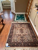 Two rugs laid out on hardwood floor showing the floral patterned larger rug in beige and dark blue with fringe and the smaller green and cream rug with floral pattern.
