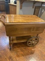 Side view of the vintage wooden rolling bar cart with drop-down side in folded position on wood floor.