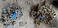Two coiled steel chains lying side by side on a concrete floor, one appears silver metallic, the other has a bronze finish.
