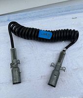 Black coiled electrical cord for transport truck with metal plug-in connectors on both ends, shown uncoiled partially.