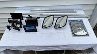 Top-down view of mirror extension kit parts on a white cloth, showing metal brackets, long and short poles, two used mirrors, and a new mirror in packaging.