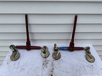 Two half ratchet load binders placed side by side on a white surface in front of white siding, showing red handles and metal hooks.