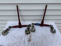 Pair of ratchet load binders with red handles and metal hooks on white surface against white siding wall.