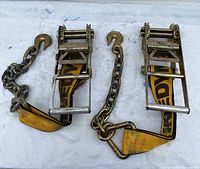 Front view of the two ratchets with attached yellow straps and hooked chains laid out on a white surface.