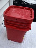 Side angle view of sealed red pail containing tire chains outside on snow near a building showing lid and handle details.