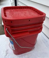 Red plastic pail with closed lid holding tire chains, shown in snowy outdoor setting.