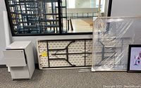 Folded plastic folding table with black metal legs, white wooden filing cabinet, framed picture wrapped in plastic leaning against wall