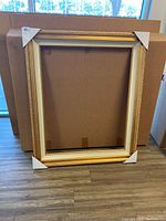 Full view of one gold gilted picture frame standing on the floor against cardboard boxes.