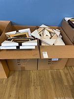 Two cardboard boxes on a wooden floor filled with multiple 9x12 burl wood picture frames featuring gold and silver trim. Frames are stacked with white foam corner protectors.