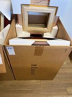 Box of photo frames showing wooden and golden trim frames stacked and protected with foam inserts inside a cardboard box.