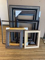 Photo showing seven various wooden picture frames stacked and leaning against a wall on a floor, with visible textures and color variations