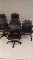 Four black leather office desk chairs placed against a wall; three chairs have swivel bases with wheels, and one is stationary without wheels.