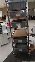 Full view of tall metal rack with five storage trays including four grey plastic tubs and one wooden platform. Wheeled base visible.