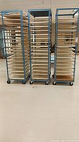 Front view of three blue metal adjustable racks with multiple cream-colored shelves each on wheeled bases.