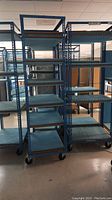 Front view of six tall blue metal shelving racks on wheels, showing the shelves and frame details.