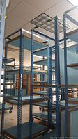 Side and perspective views of multiple tall blue metal shelving racks with ribbed shelves on caster wheels.