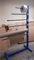 Blue metal packaging paper dispenser with several horizontal arms holding rolls of plastic wrap and kraft paper