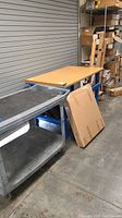 Work bench table with wooden top and blue metal legs next to plastic cart on wheels.