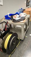 Shows two grey plastic utility carts on wheels with various items on top and below, including office supplies, paper, and plastic wrap. In front is a large black and yellow strapping reel on metal frame.
