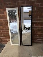 Front view showing two full length mirrors standing side by side. The left mirror has a white washed barnwood look frame and the right mirror has a black plastic frame.