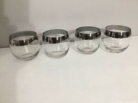Four clear roly poly style rock glasses with a thick silver rim on a white surface.