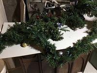 Two green Christmas garlands placed on a white surface showing their length and pine needle detail with some ornaments visible beneath.