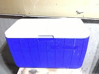 Closed blue Coleman cooler with white lid against a wall showing surface marks and dirt.
