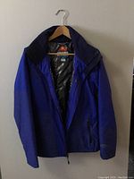 Front view of cobalt blue Columbia jacket hanging on wooden hanger on white wall showing zipper partially open, front pockets, and collar.