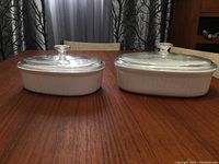 Two white CorningWare covered bakers with clear glass lids on wood table, one smaller (1.8 qt) and one larger (2.3 qt).