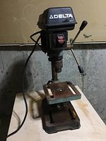 Front view of Delta drill press showing motor housing, Delta logo, work table with slots, three-handle feed mechanism, and base plate.